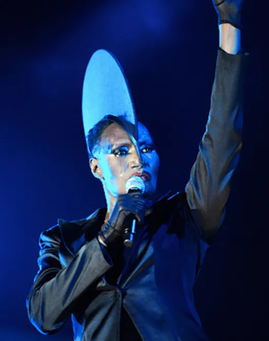 Grace Jones performing live in a towering sculptural hat, lit in electric blue